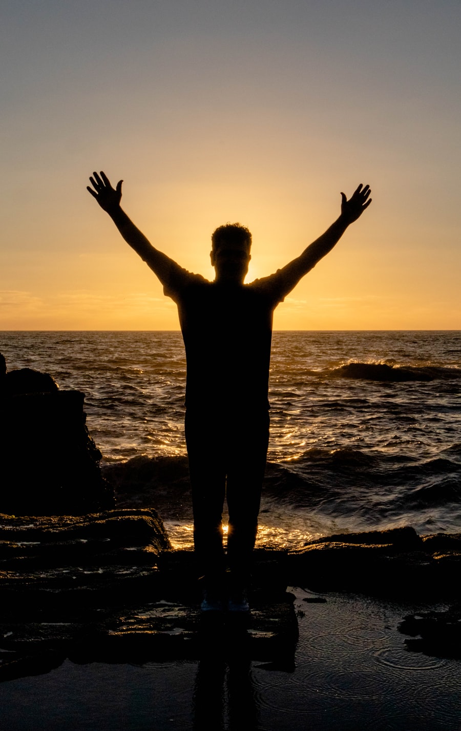 A figure at the ocean&rsquo;s edge with arms lifted toward the sunset